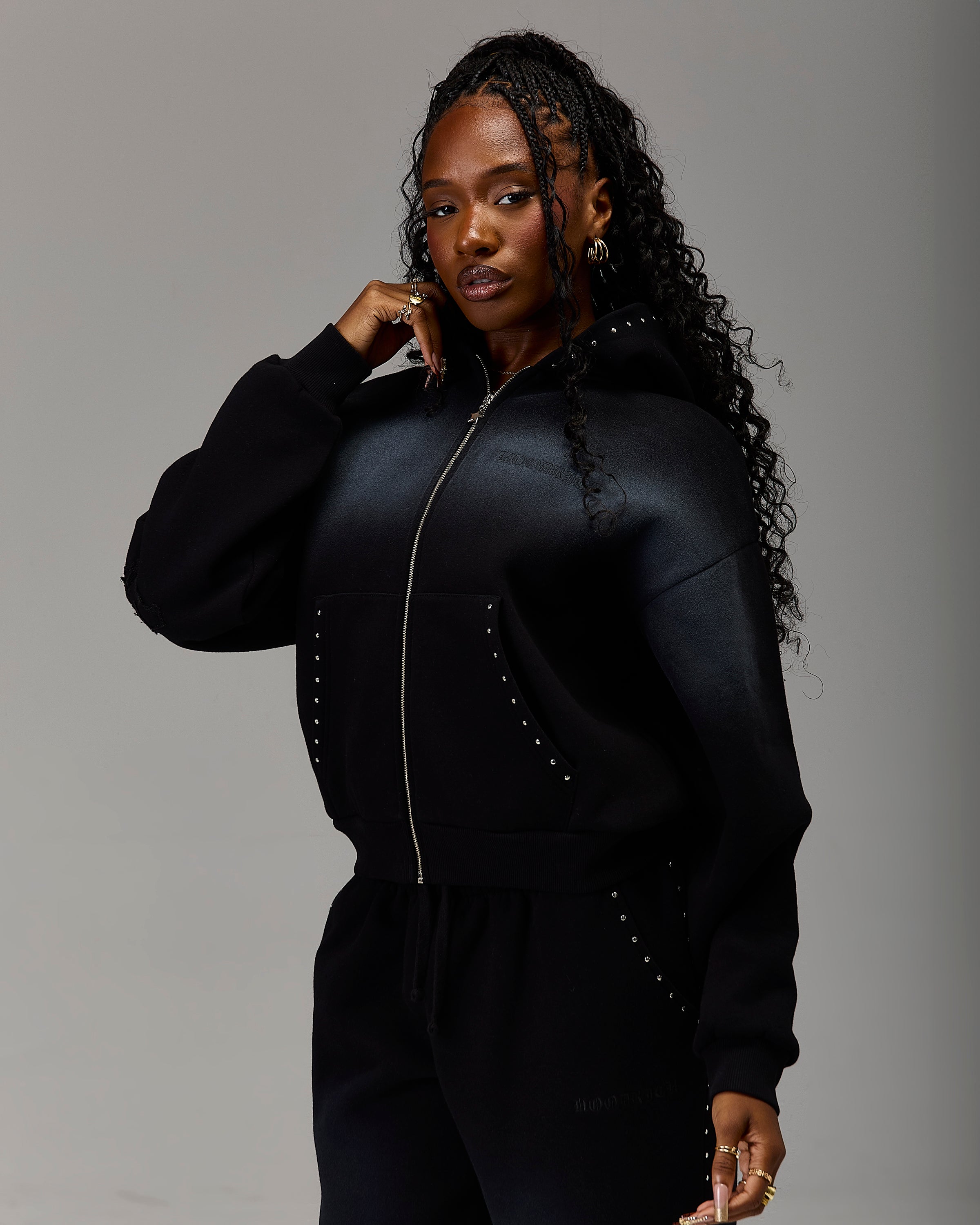 A woman modeling a black zip-up hoodie and matching sweatpants set against a plain gray studio background. The hoodie features a shiny black satin-like finish with silver stud embellishments along the front pockets and zipper area.