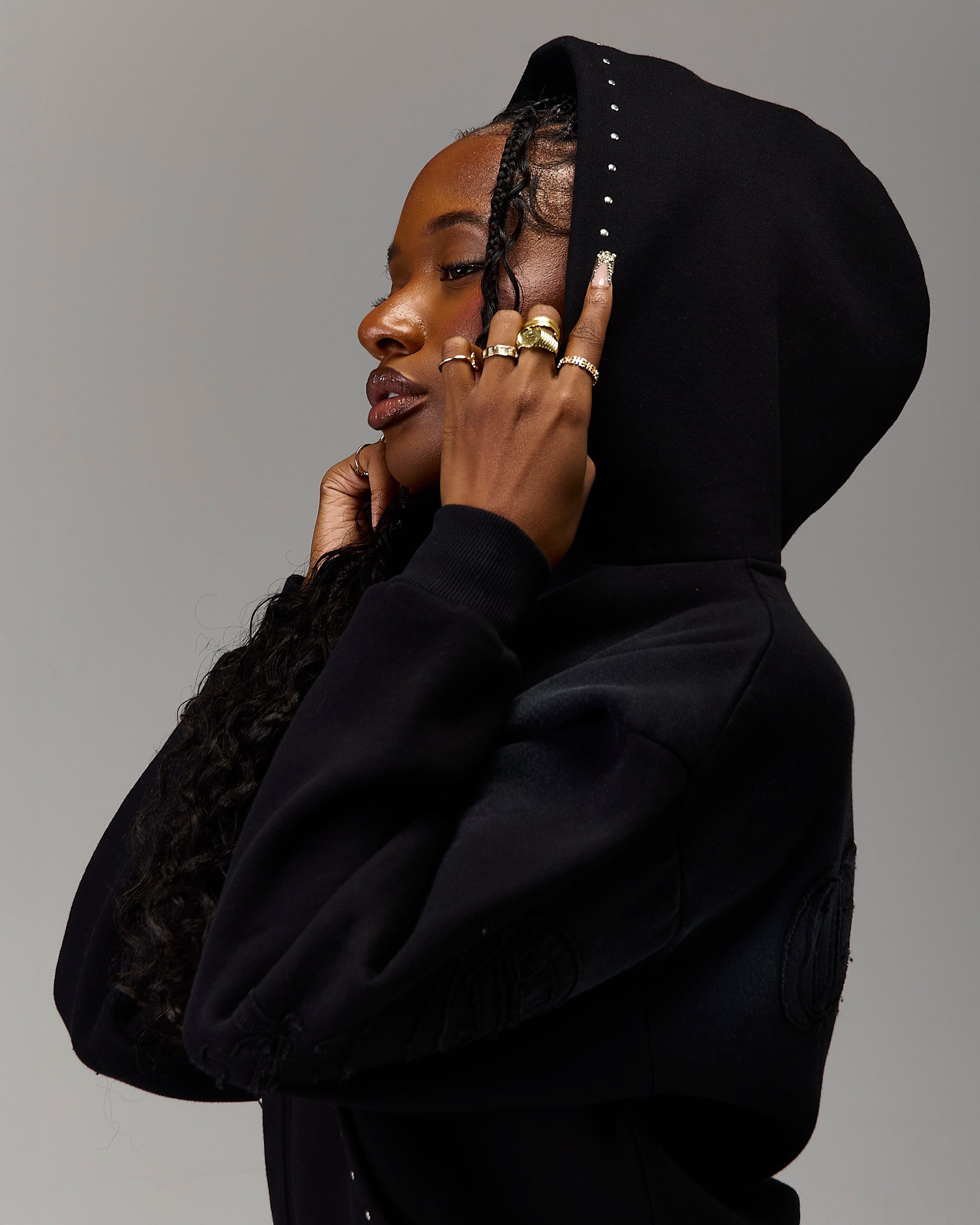 A woman in profile wearing a black hooded sweatshirt with silver studs along the hood edge, posed against a solid gray studio background.