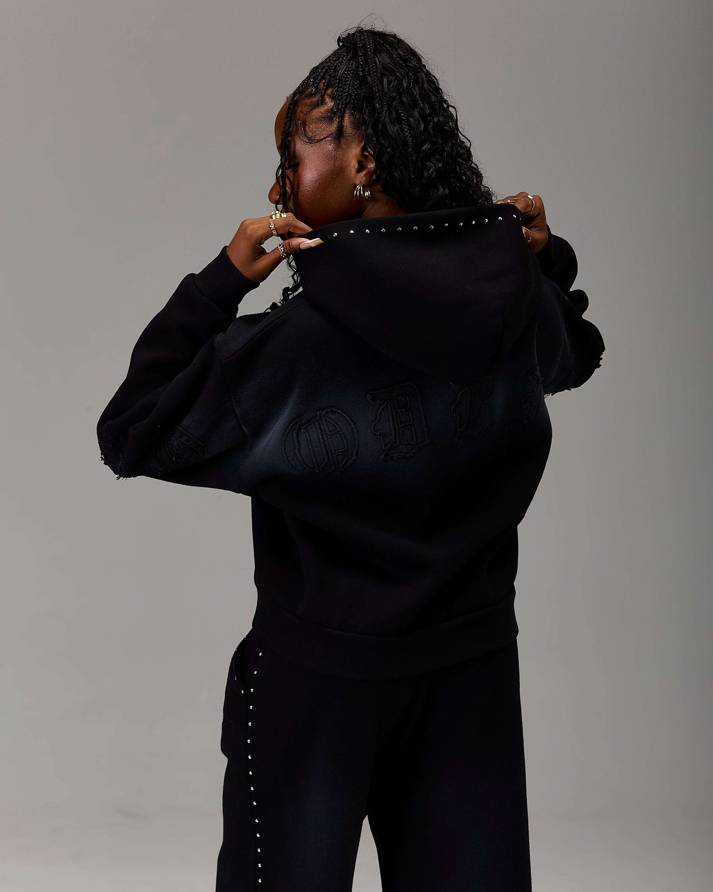 A model wearing a black hoodie and matching black pants, viewed from behind as she holds up the hood of the sweatshirt. The hoodie features tonal black embroidery of the Greek letters Hoodrich on the back, with silver stud detailing along the collar and down the side of the pants.