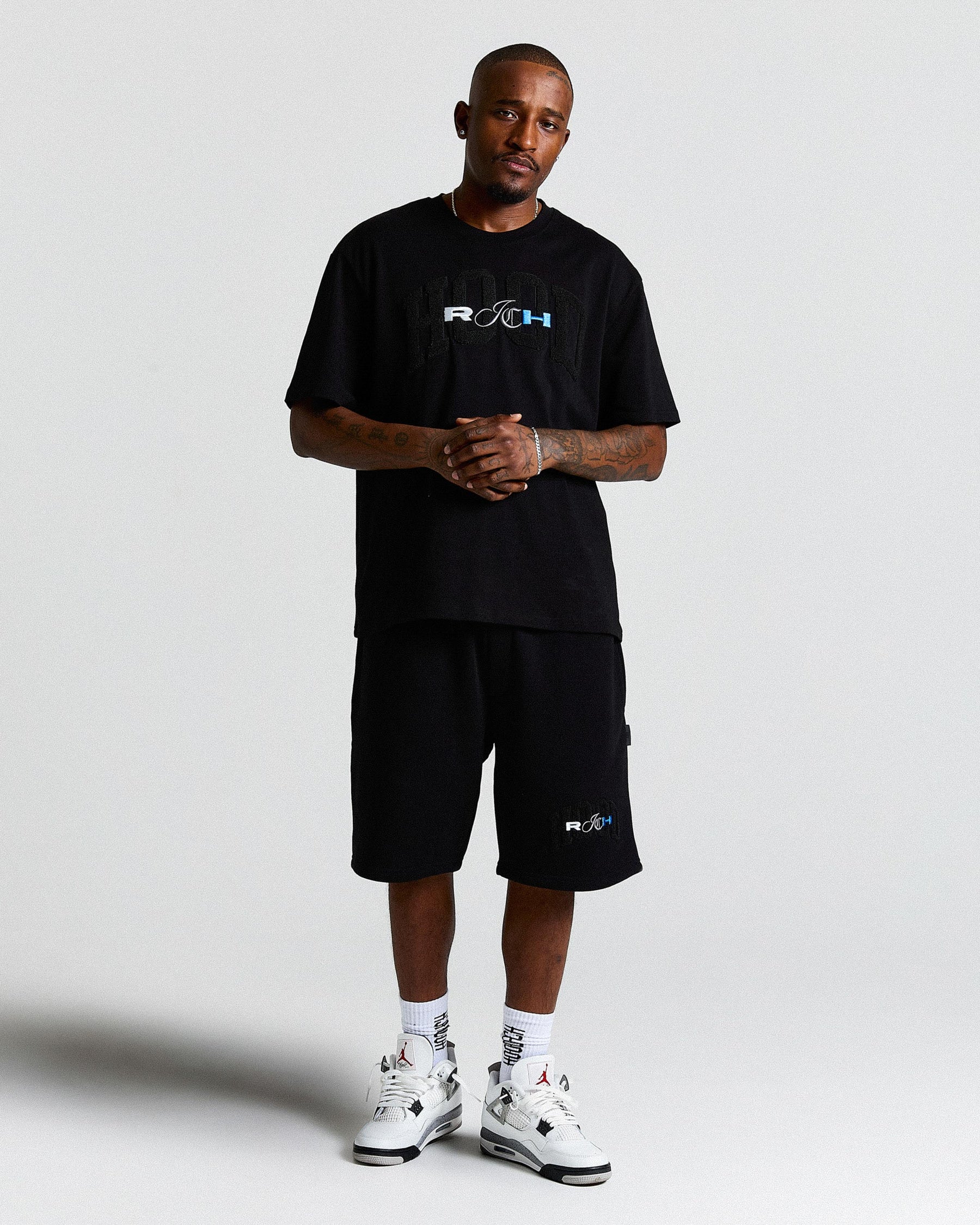 Man wearing a black t-shirt and shorts with logos, standing against a white background