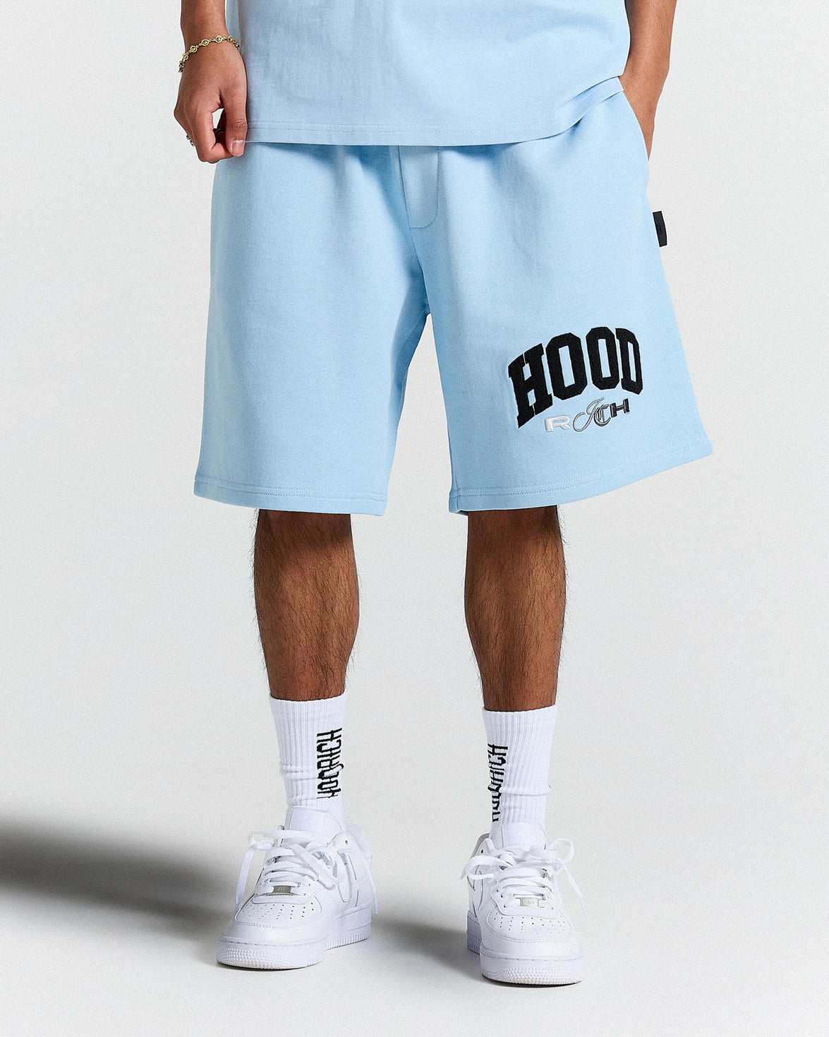 Person wearing light blue shorts with 'HOODRICH' print, white socks, and white sneakers on a light grey background