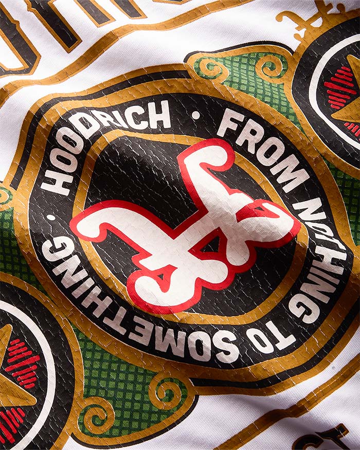 Close-up of a graphic print featuring the brand name 'HOODRICH' and their signature red stylized logo on a black circular badge, surrounded by gold and green decorative borders on a white background.