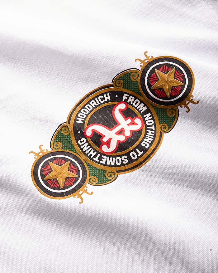 A close-up of a graphic print on white fabric, featuring the band Hoodrich's logo with the text 'FROM NOTHING TO SOMETHING'. The design includes a central black oval with a red and white stylized 'HX' logo, surrounded by green and gold decorative elements, and two circular gold star motifs on either side.