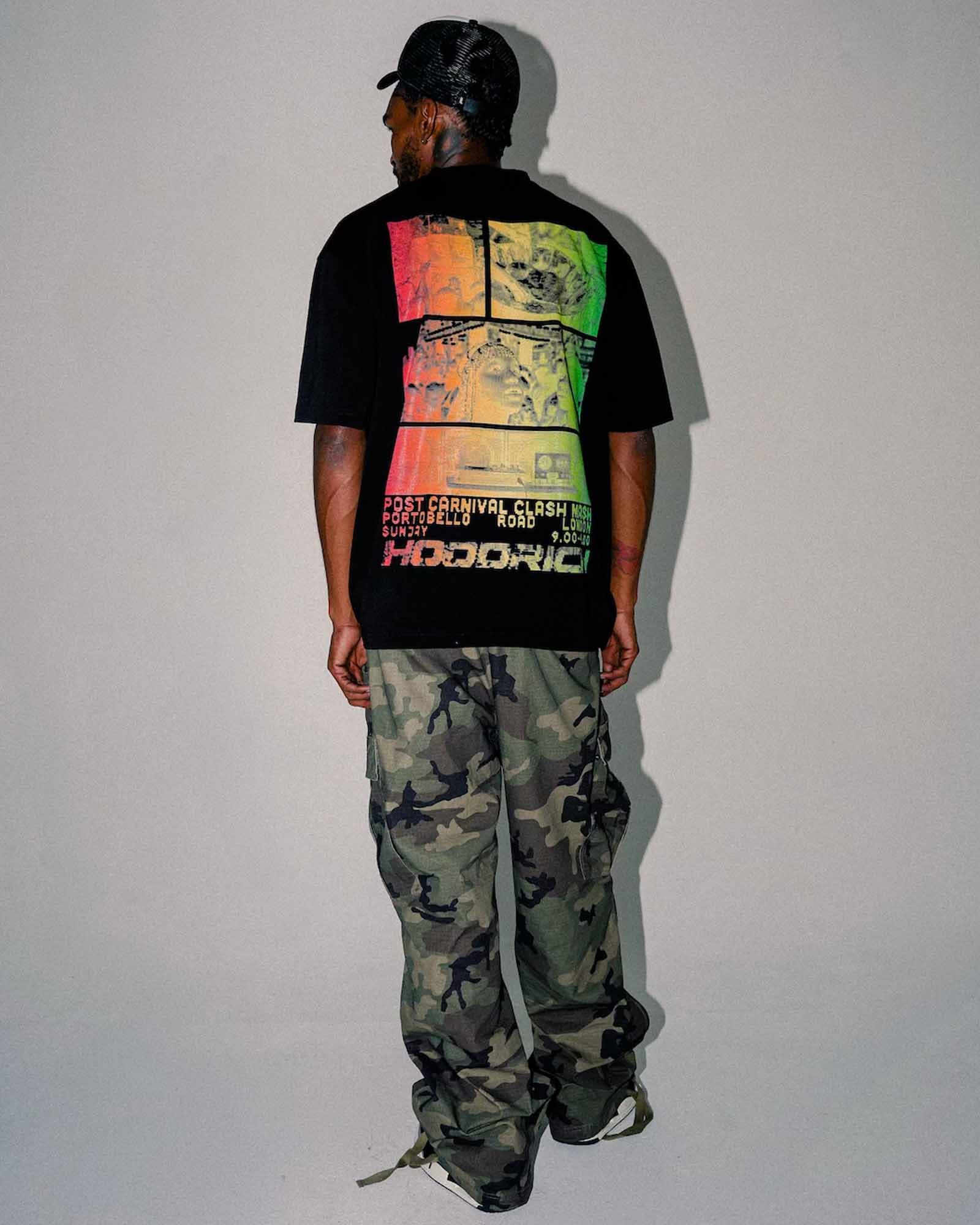 A man viewed from behind wearing a black HODORICH t-shirt with a large multicolor graphic print on the back, paired with green and brown camouflage cargo pants, standing against a plain light gray wall.
