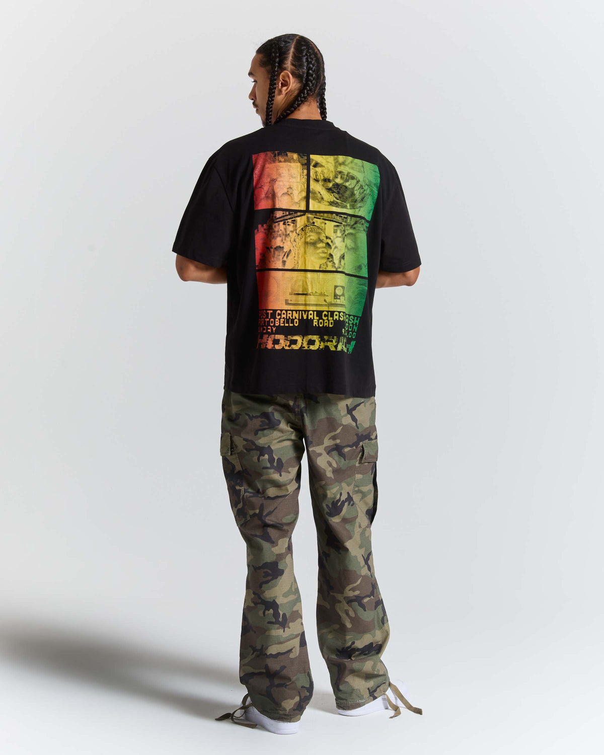 A man viewed from behind wearing a black t-shirt with a large multicolor graphic print on the back, paired with green and brown camouflage cargo pants, against a plain light gray background.