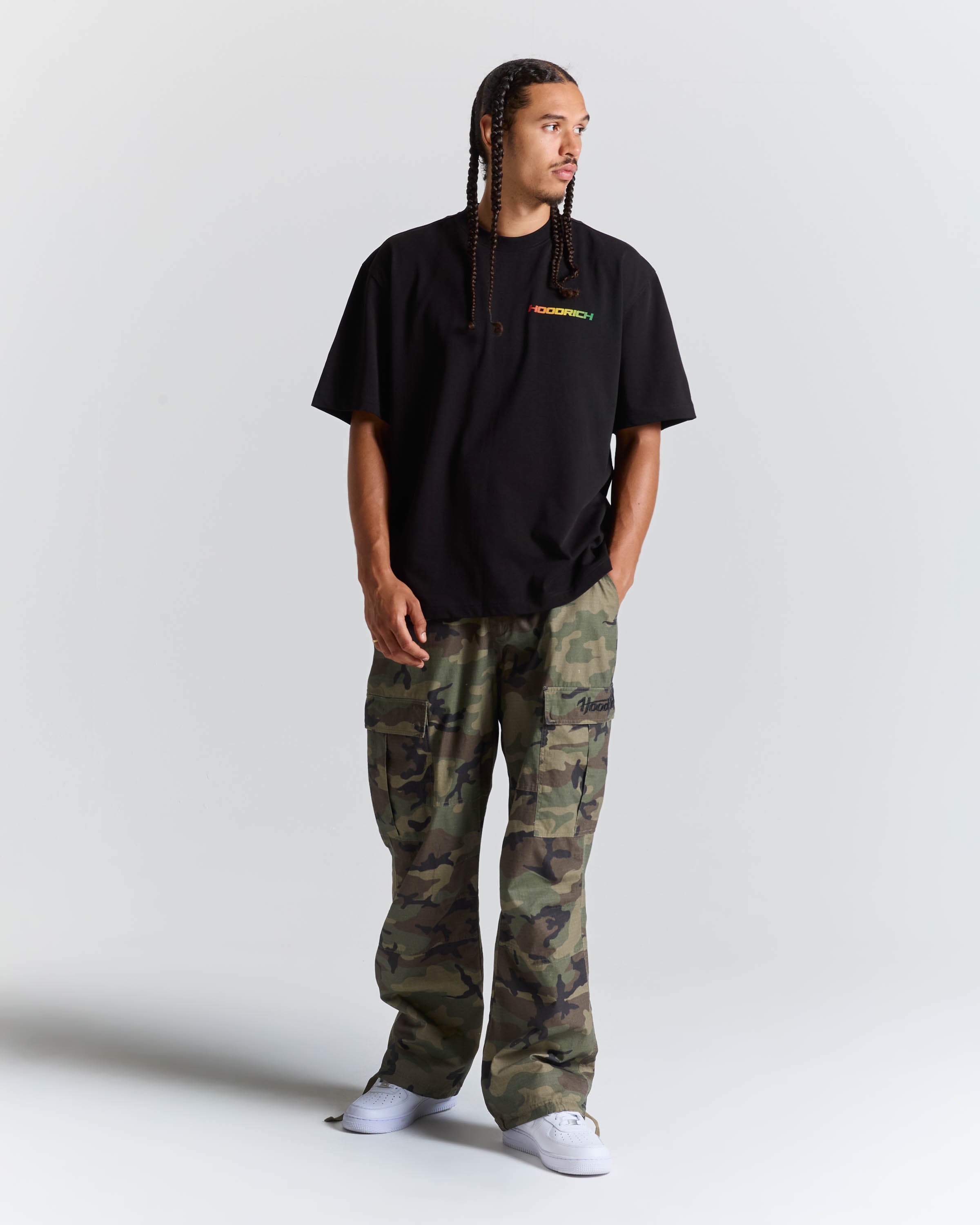 A man modeling a black t-shirt with a small green and yellow 'HOODRICH' logo on the chest, paired with green and brown camouflage cargo pants and white sneakers, against a light gray studio background.