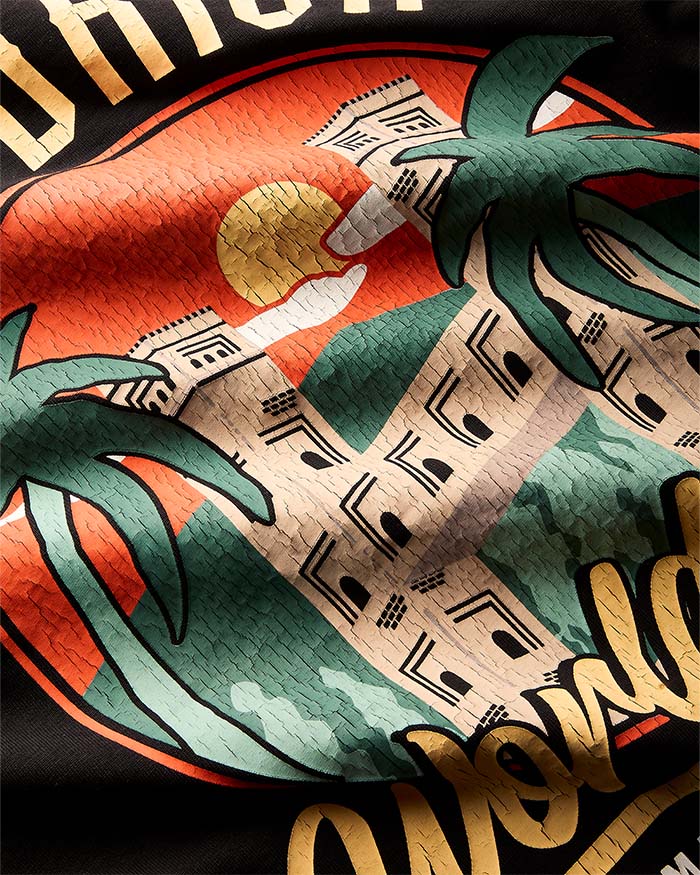 Close-up of a graphic print featuring a stylized desert scene with a beige building, green palm trees, a red-orange sun, and gold lettering on a dark background.
