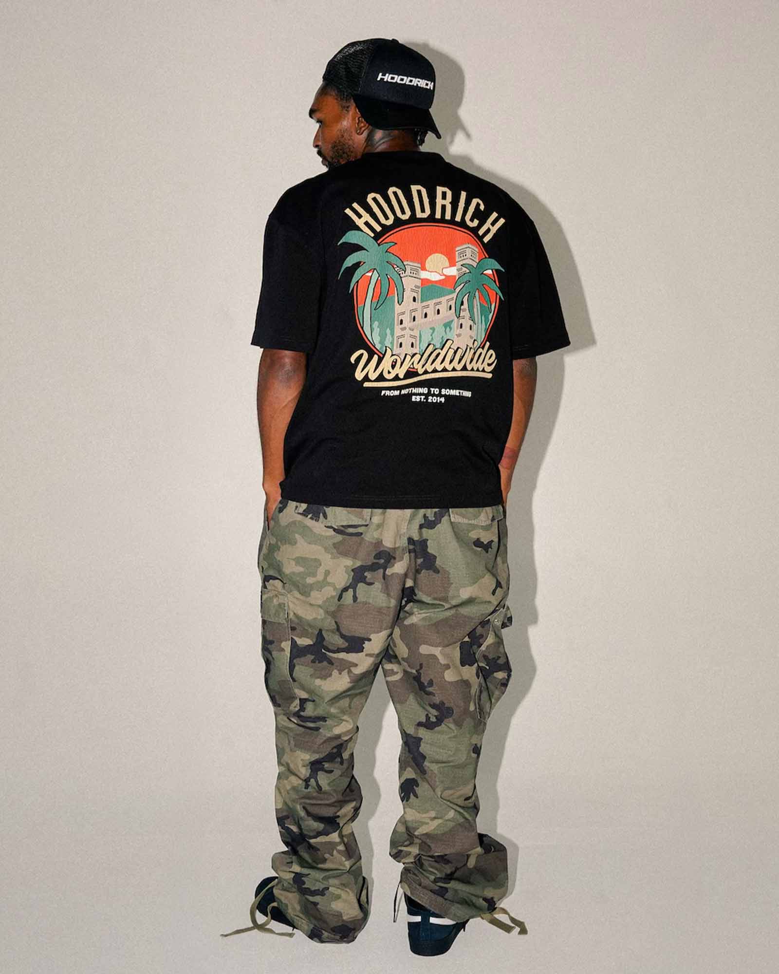 A man shown from the back wearing a black HOODRICH t-shirt with a large colorful graphic on the back, paired with green and brown camouflage cargo pants, standing against a plain light gray wall.
