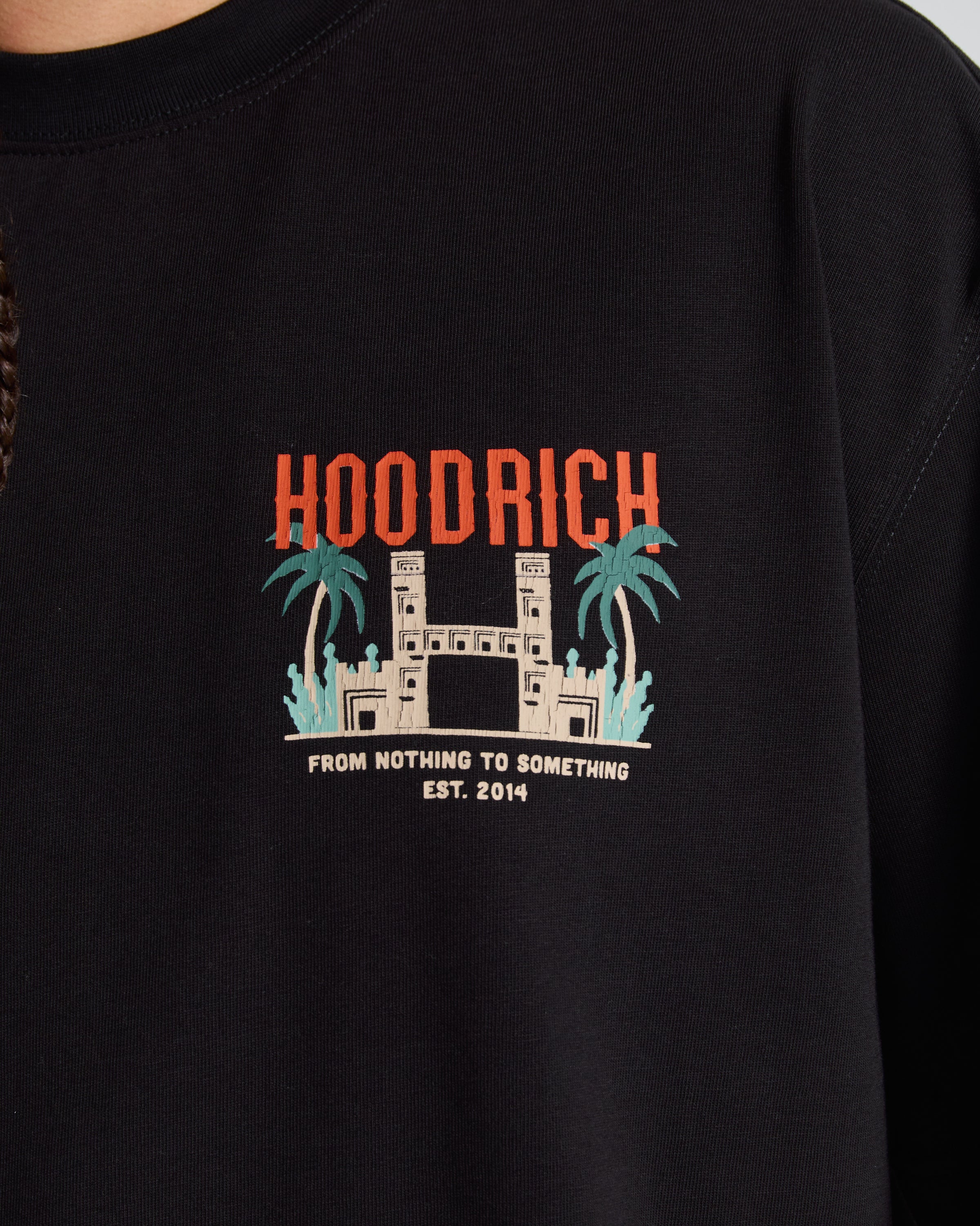 Close-up of a black crewneck sweatshirt featuring a graphic print on the chest with the word 'HOODRICH' in orange, a white gate illustration with teal palm trees, and the text 'FROM NOTHING TO SOMETHING EST. 2014'.