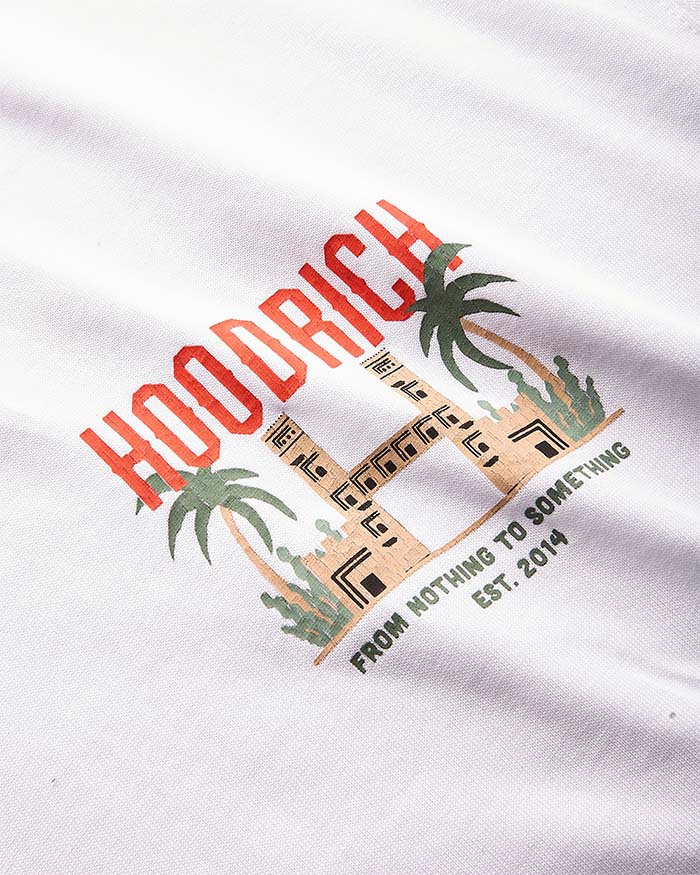 A close-up of a white fabric surface featuring a printed graphic with the word 'HOODRICH' in red-orange letters, palm trees, a building illustration, and the phrase 'FROM NOTHING TO SOMETHING EST. 2014'.