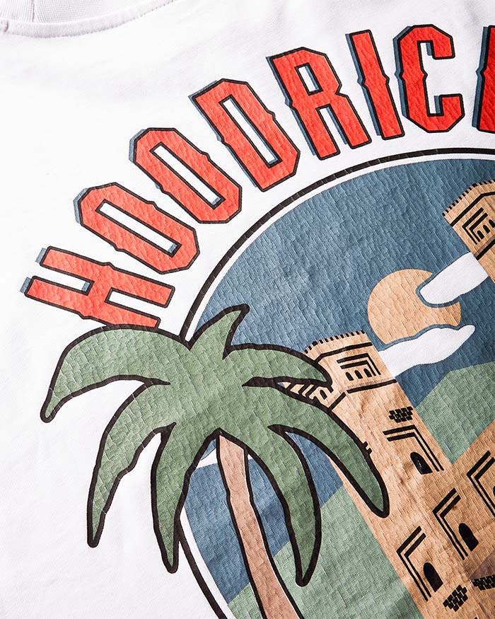 Close-up of a white graphic t-shirt featuring the word 'HOODRICH' in bold red letters above a circular illustration with a green palm tree, blue sky, and tan buildings.
