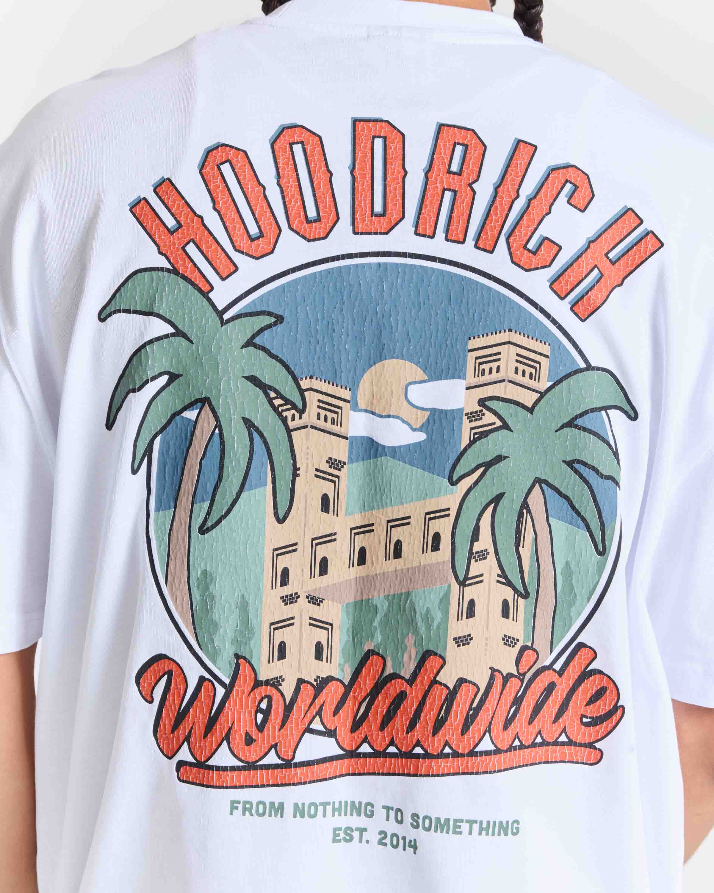 White short-sleeve graphic T-shirt shown from the back on a model, featuring a large embroidered-style design with the words 'HOODRICH' and 'Worldwide' in orange-red lettering, flanking a circular illustration of palm trees, a building, and a sun.
