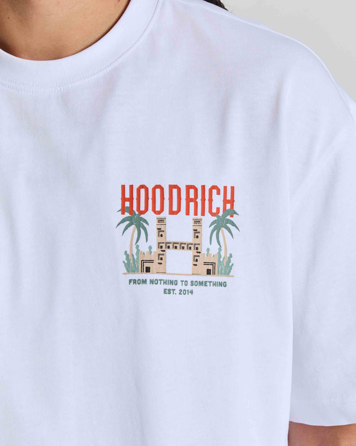 A close-up of a person wearing a white crewneck t-shirt with a colorful graphic on the chest. The graphic features the word 'HOODRICH' in red-orange letters above a beige building flanked by green palm trees, with the text 'FROM NOTHING TO SOMETHING EST.
