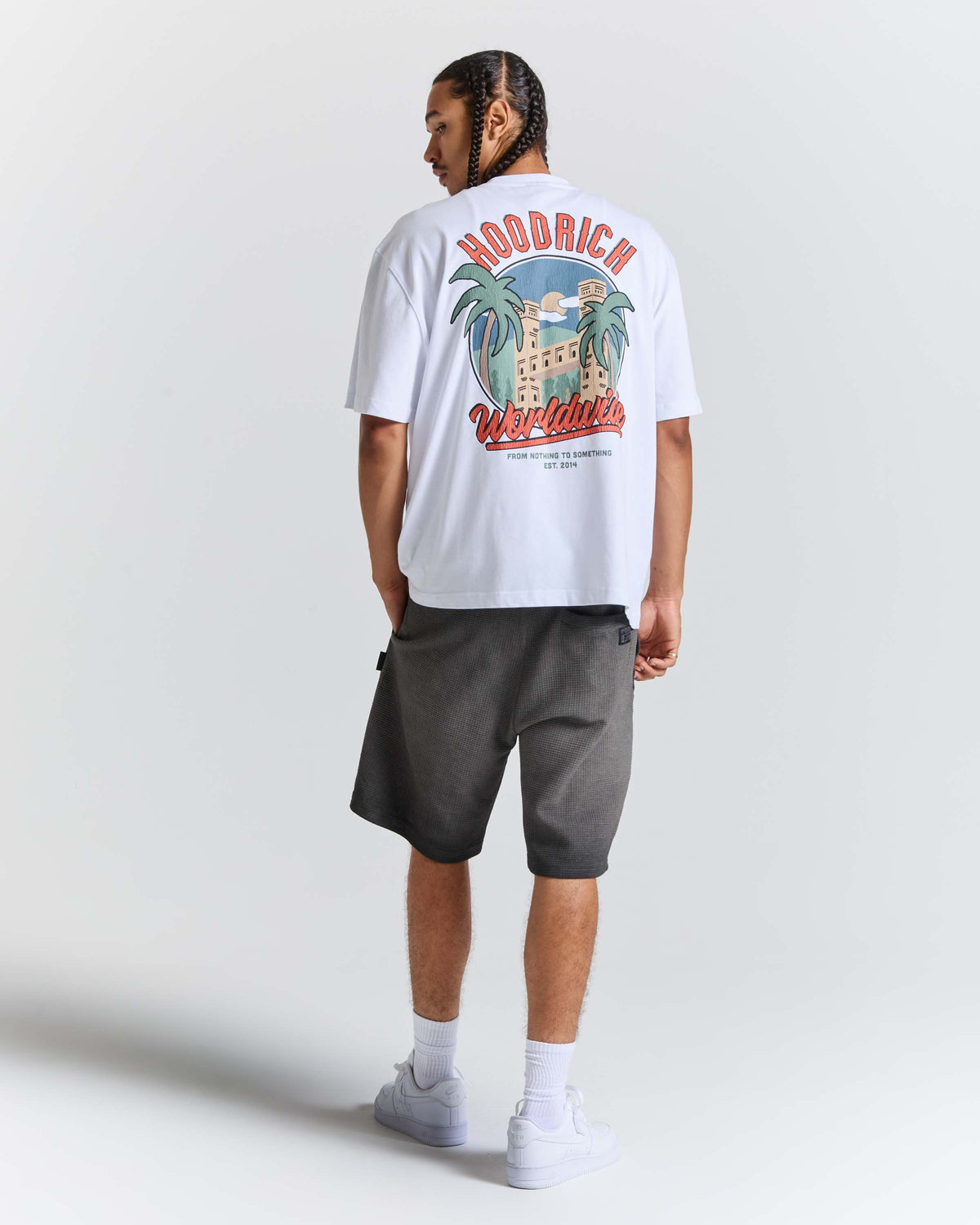 A man wearing a white HODRICH t-shirt with a large colorful graphic on the back, paired with dark gray shorts, white socks, and white sneakers, shown from a rear three-quarter view against a light gray studio background.