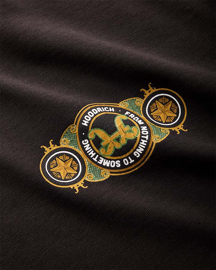 A close-up of a black fabric garment featuring a decorative embroidered patch. The patch has a gold border, green textured details, and a central green snake motif with the text 'HOODRICH FROM NOTHING TO SOMETHING' in white.