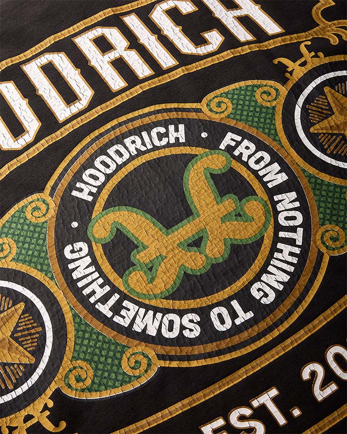 Close-up of a graphic print featuring the word 'HOODRICH' in large white letters, a central circular emblem with a stylized green and gold symbol, and decorative gold and green scrollwork on a black background.