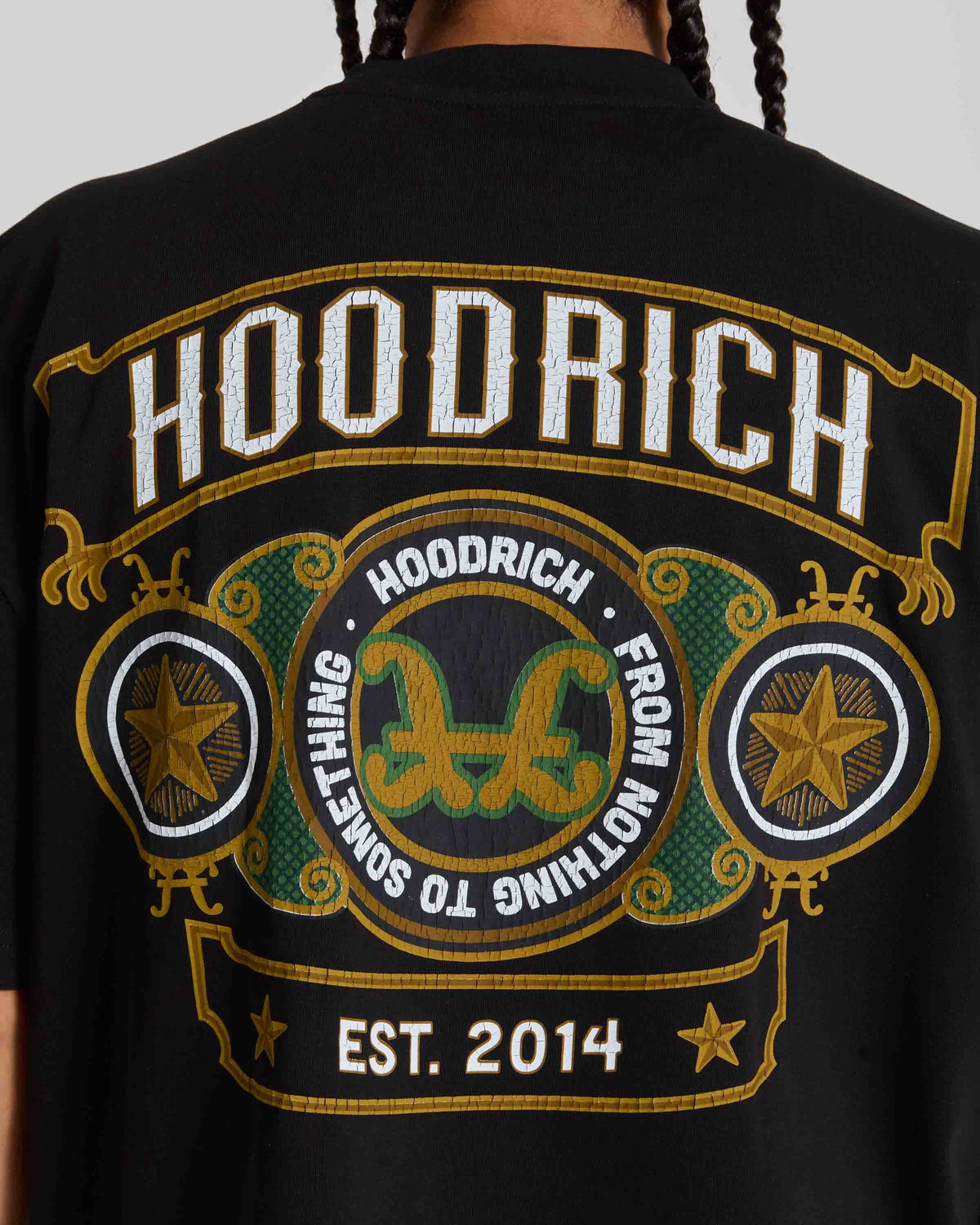 Close-up of the back of a black hoodie featuring a large embroidered graphic with the brand name 'HOODRICH' in white outlined letters, gold borders, green and gold decorative elements, and 'EST. 2014' near the bottom.