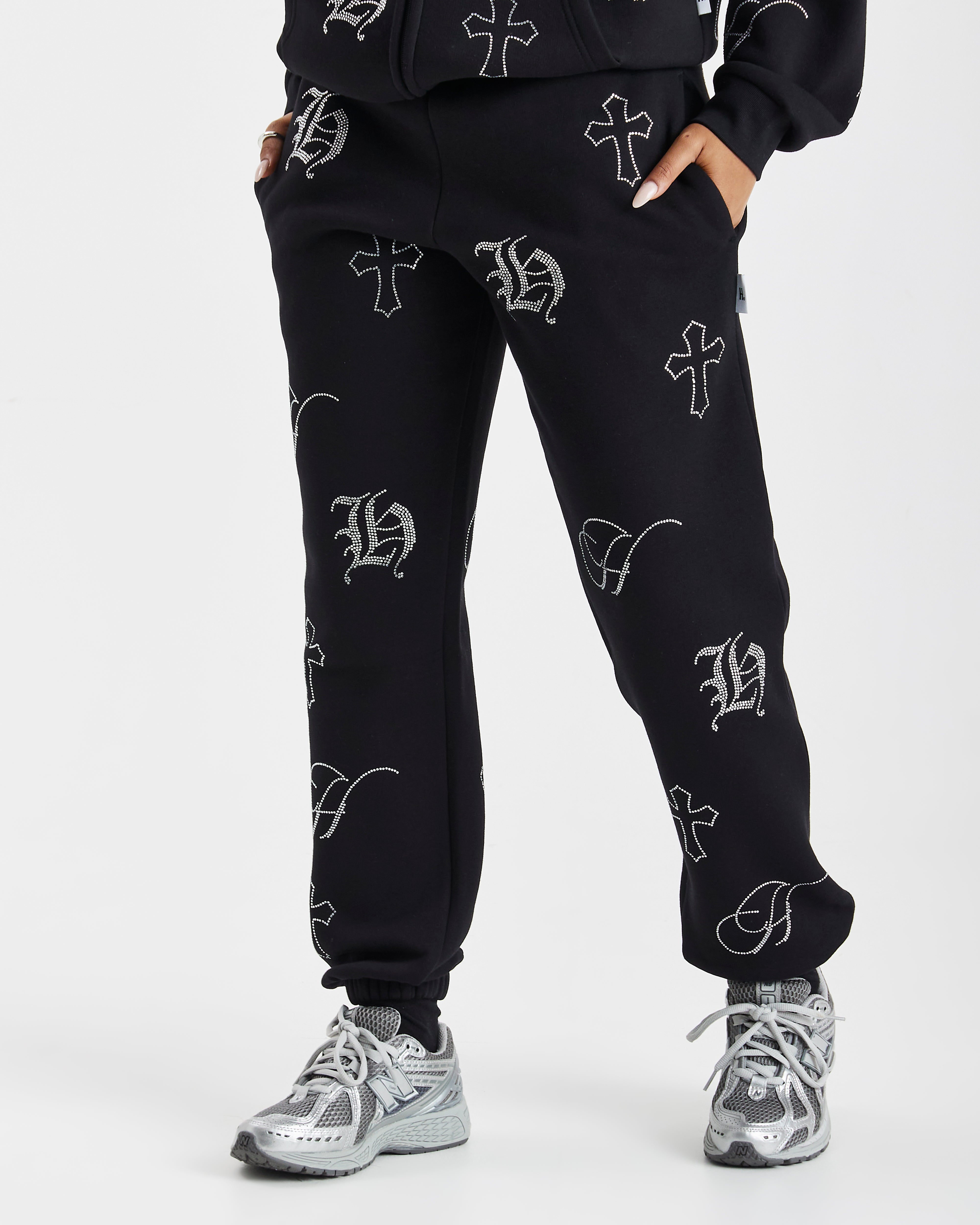 Black sweatpants with an all-over white Gothic monogram print, shown on a model wearing gray and white sneakers against a light gray studio background.
