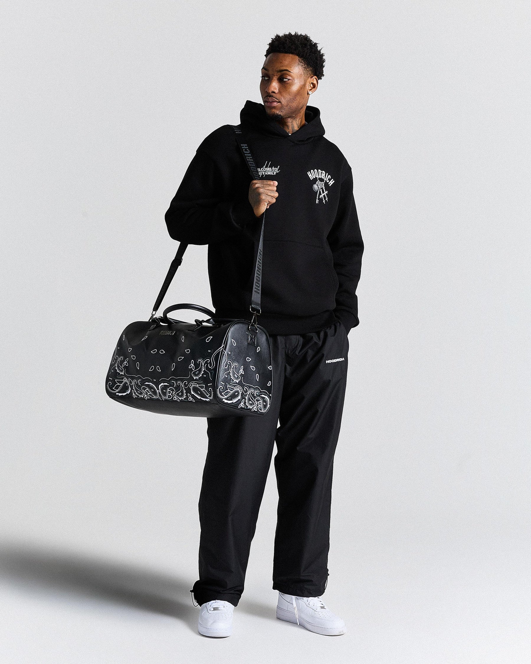 Person wearing a black hoodie and pants holding a black duffel bag with white patterns on a light gray background
