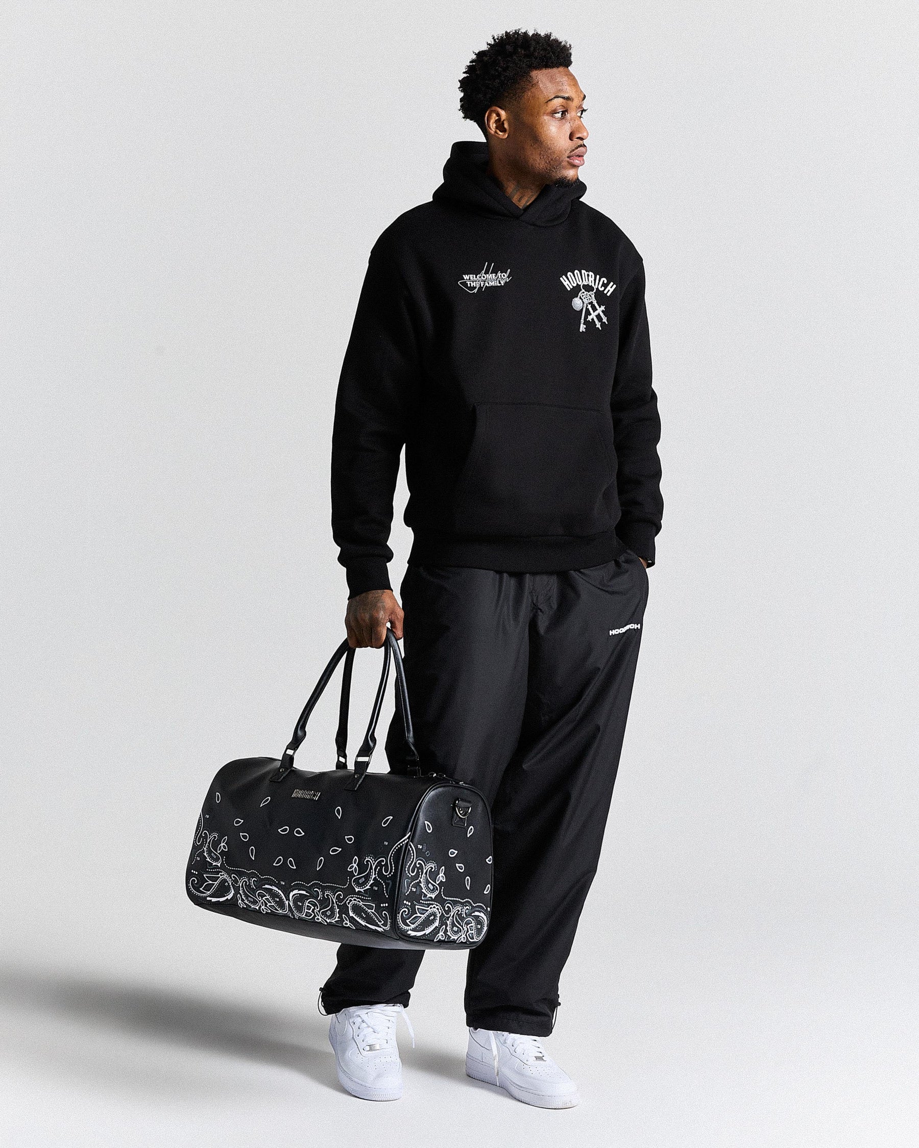 Man wearing a black hoodie and pants holding a black duffel bag with white patterns on a light gray background