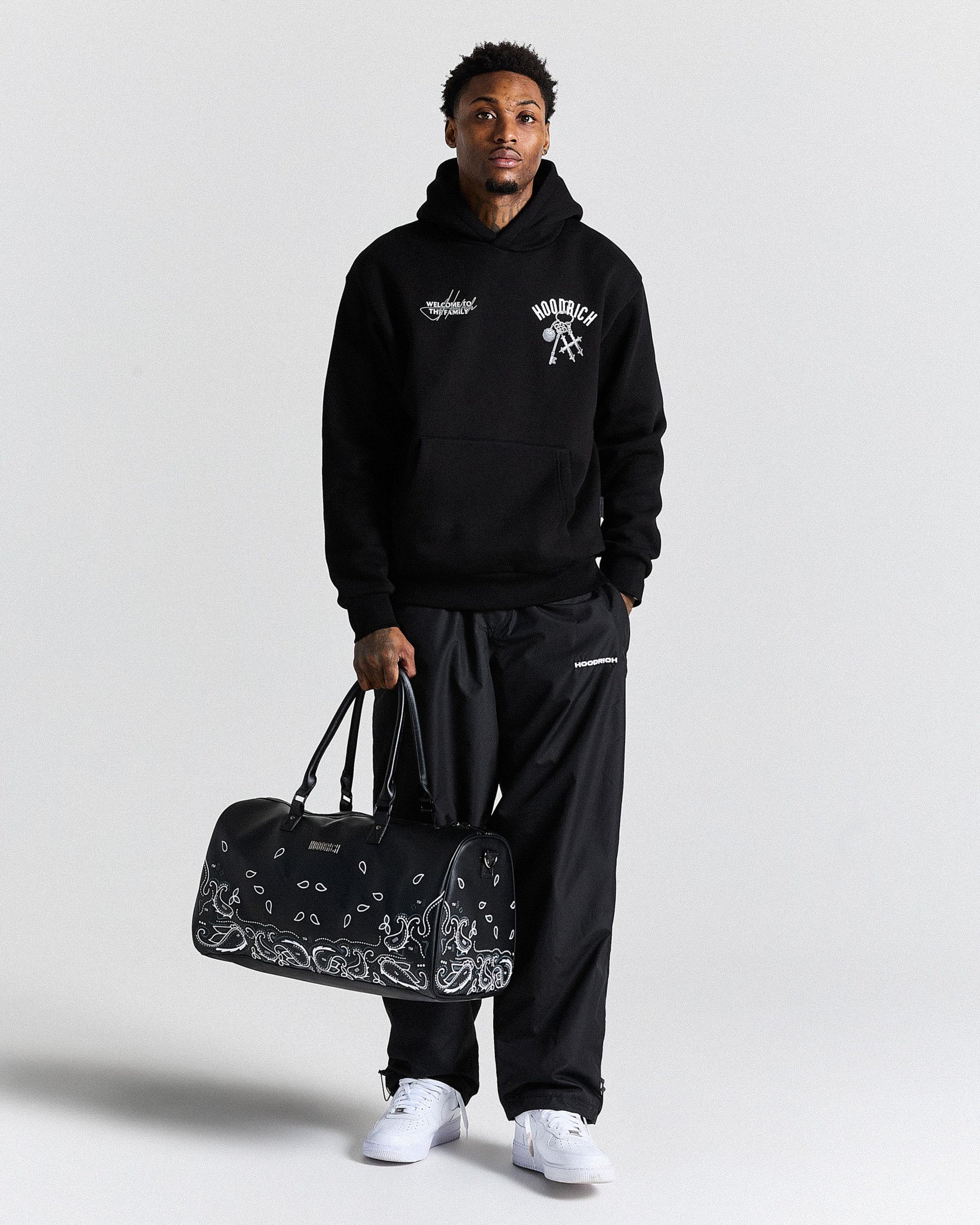 Person wearing a black hoodie and pants holding a black duffel bag with bandana design on a white background