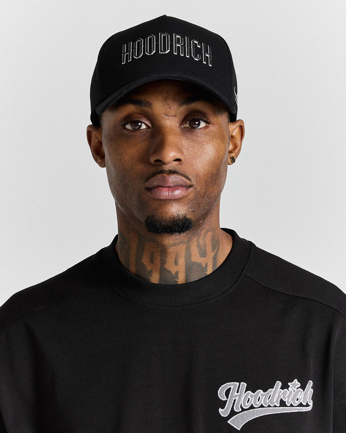 Man wearing a black 'Hoodrich' cap and sweatshirt against a gray background