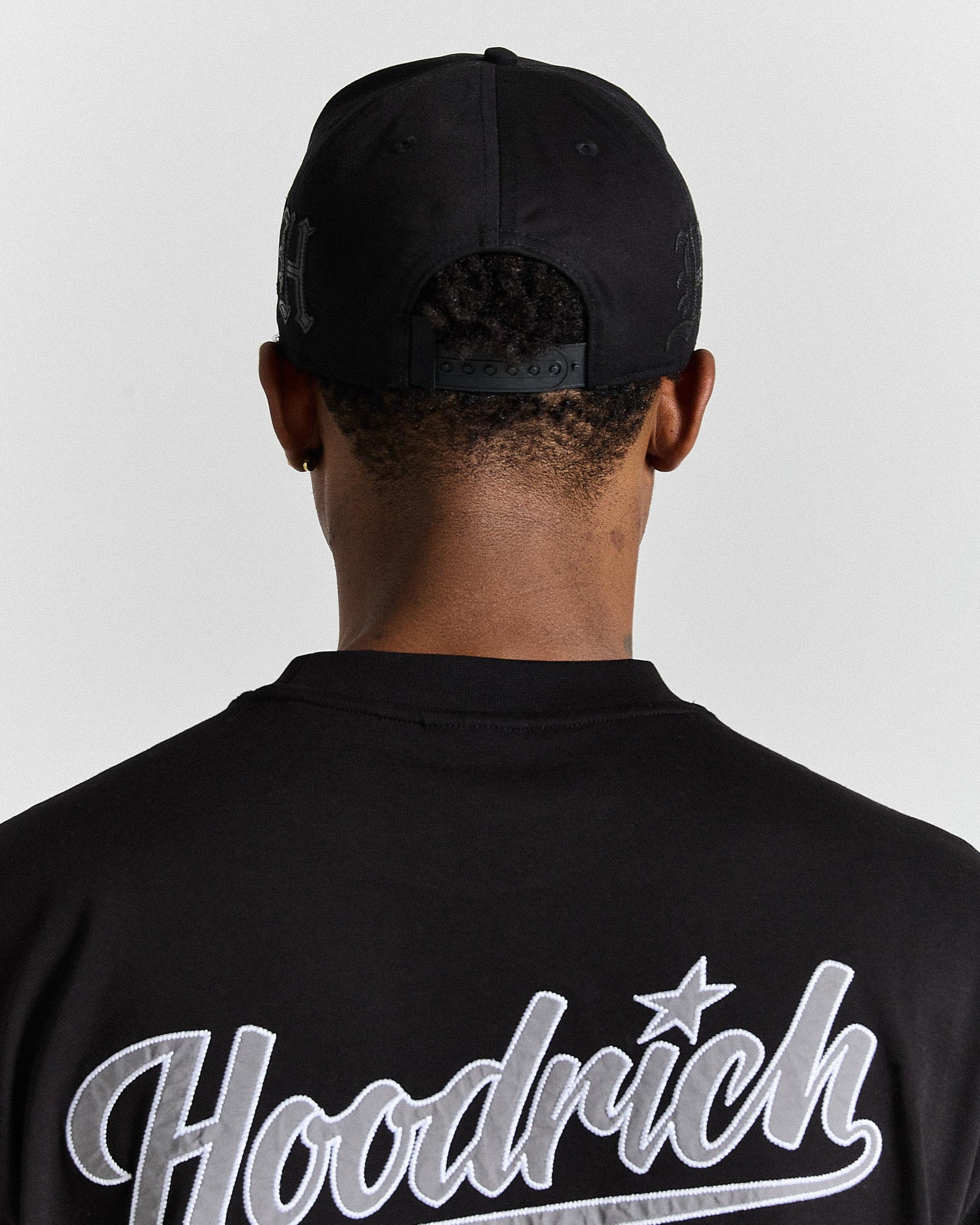 Person wearing a black cap and black 'Hoodrich' t-shirt on a light gray background