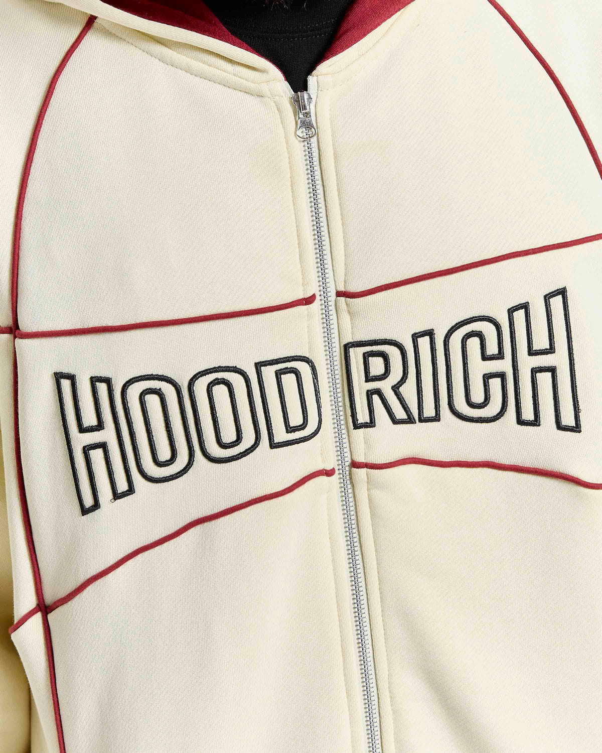 Close-up of a cream zip-up hoodie with 'HOOD RICH' printed across the chest in black outlined letters, accented by dark red piping and a silver zipper.