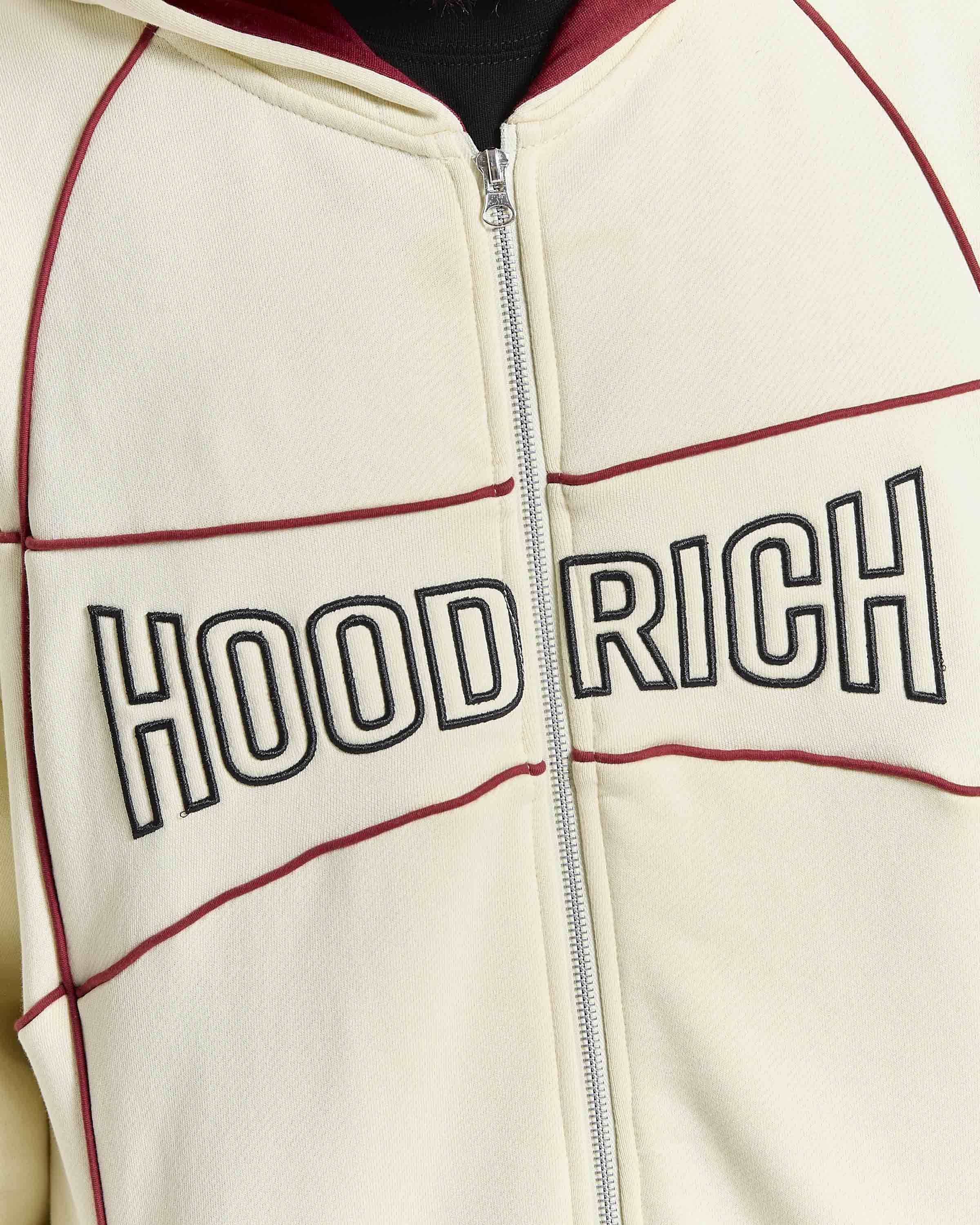 Close-up of a cream zip-up hoodie with 'HOODRICH' printed across the chest in black outlined letters, accented by dark red piping and a silver zipper.