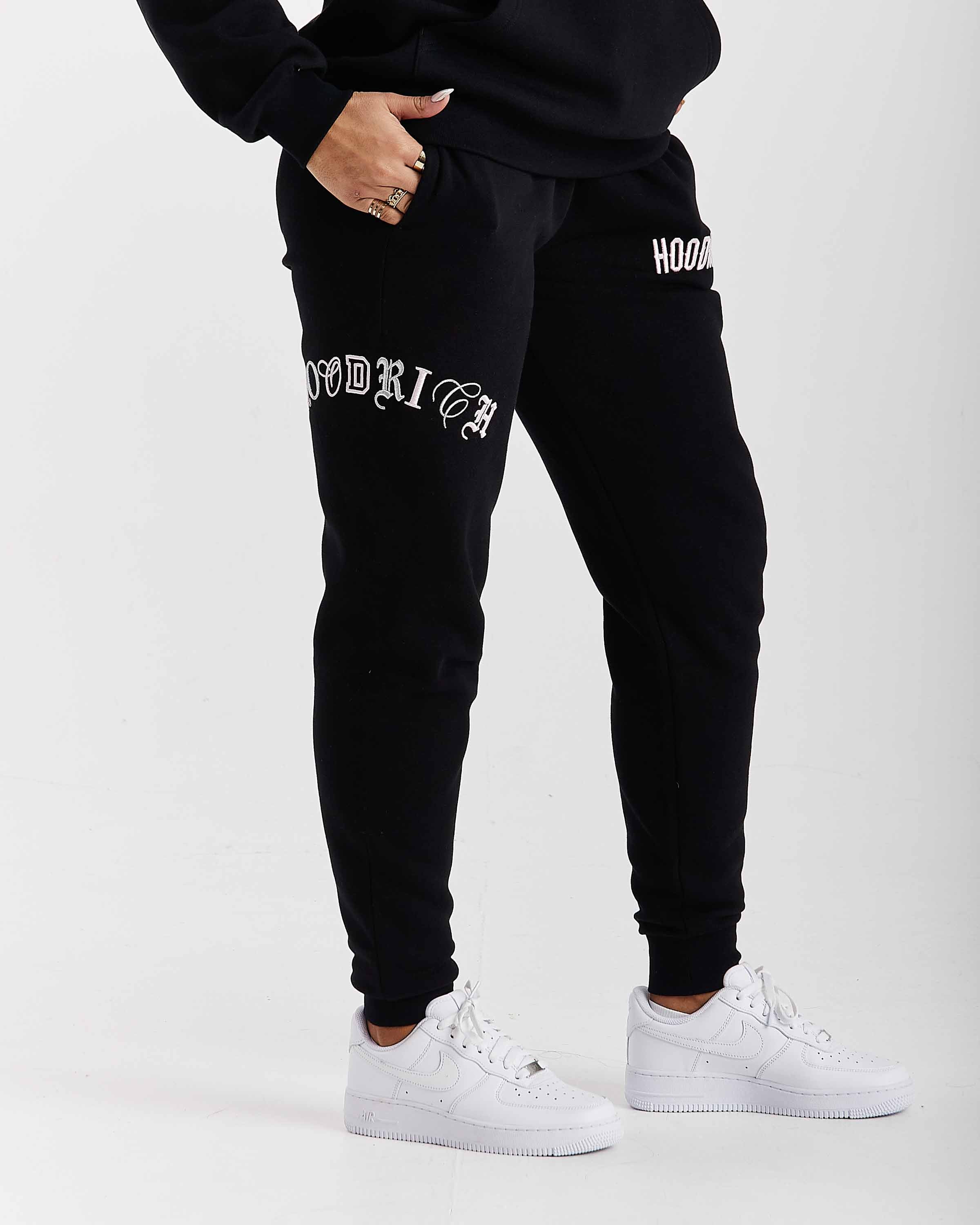 Person wearing black jogger pants with white text and white sneakers on a light gray background