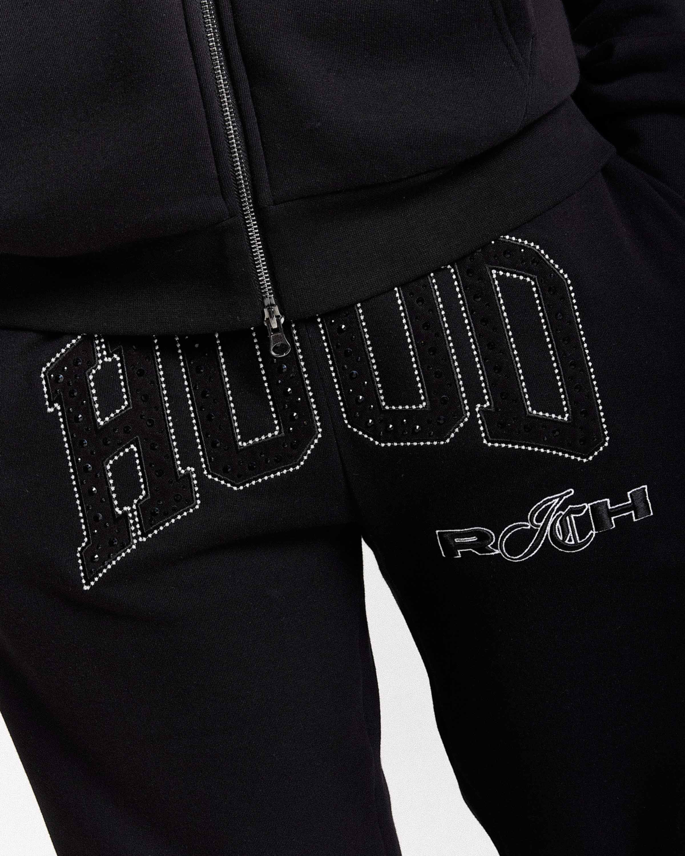 Close-up of black sweatpants with large rhinestone-studded 'Hood' lettering across the thigh and a white 'Rich' logo below, paired with a black zip-up top.