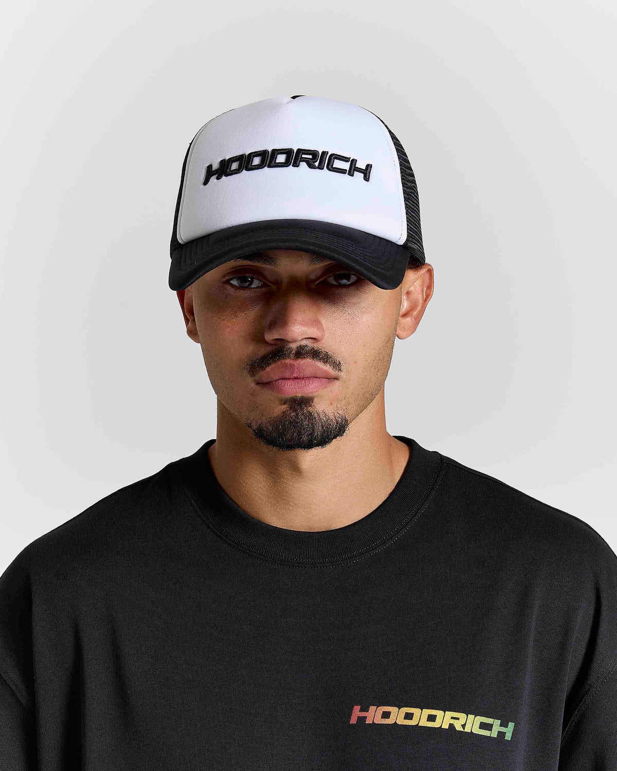 A man wearing a black and white trucker hat with 'HOODRICH' printed on the front, paired with a black crewneck sweatshirt featuring a small multicolor 'HOODRICH' logo on the chest, set against a light gray background.