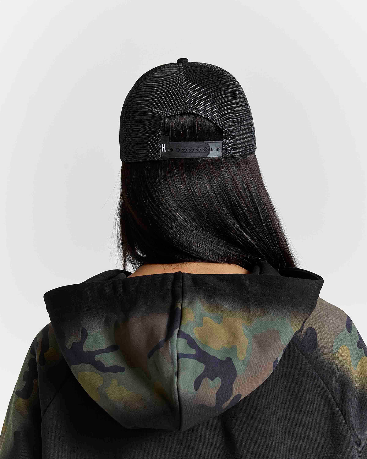 Back view of a person wearing a black trucker cap and a black hoodie with a green and brown camouflage pattern on the hood, set against a light gray background.