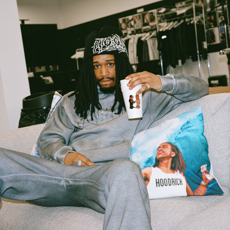 A man wearing a gray sweatshirt and matching sweatpants sits on a light gray couch in a clothing store, holding a white mug. A square pillow with a printed portrait and the word 'HOODRICH' rests beside him.