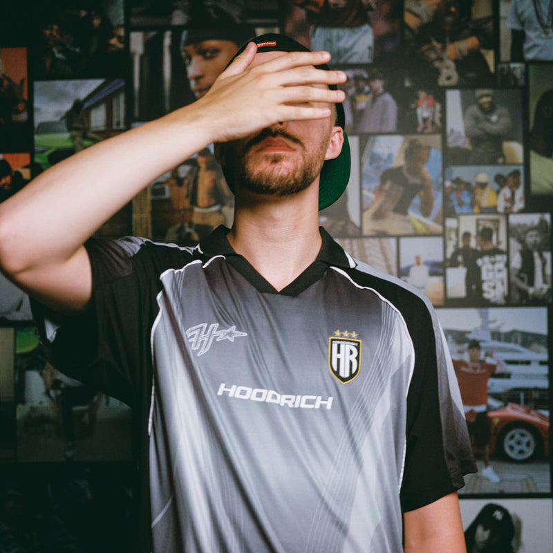 A man wearing a gray and black HOODRICH sports jersey, covering his eyes with his hand, set against a dark background of framed photos.